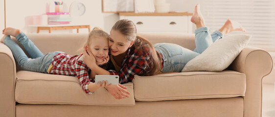 Happy mother and little daughter with smartphone resting on sofa at home. Banner design