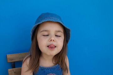 A girl in a Panama hat grimaces and shows her tongue against a blue isolated background