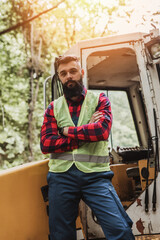 Young adult lumberjack or logger standing in front of huge bagger excavator in the woods and looking in the camera with crossed arms.