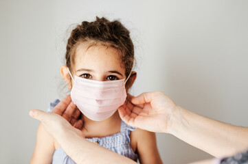 Girl with coronavirus mask prepared to go outside. Mother's hands putting the mask on her girl to prevent contagion