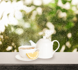 Teapot and cup of hot tea with lemon near window on rainy day