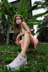 Young beautiful girl in pink skirt wearing fancy nike sport sneakers in the against coronovirus mask near palm tree in Thailand - obrazy, fototapety, plakaty
