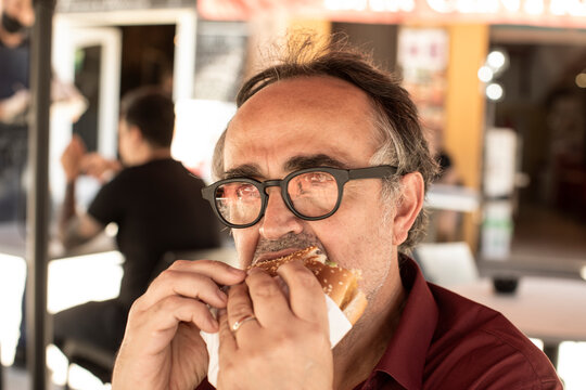 Uomo con occhiali mangia un panino con cotoletta seduto al tavolino di un bar