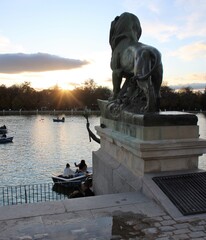 Personas navegando en bote con remos en el parque del Retiro al atardecer con una estatua de un león a la vista