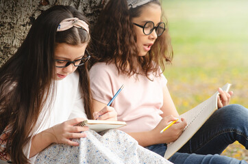 Two girl friends sitting at the base of big tree trunks and writing a book in the park in the summer