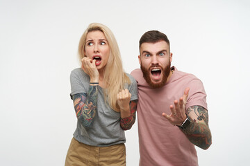 Excited young handsome brunette tattooed guy screaming excitedly and raising his hand while posing over white background with scared pretty blonde female in grey t-shirt