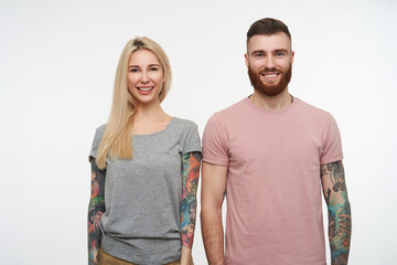 Fototapeta premium Studio photo of young lovely cheerful tattooed couple looking positively at camera with wide smiles and keeping their hands along bodies, posing over white background