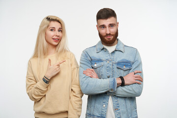 Studio photo of young pretty blonde female in beige sweatshirt pointing positively on cute young brown haired bearded man with folded hands, posing over white background