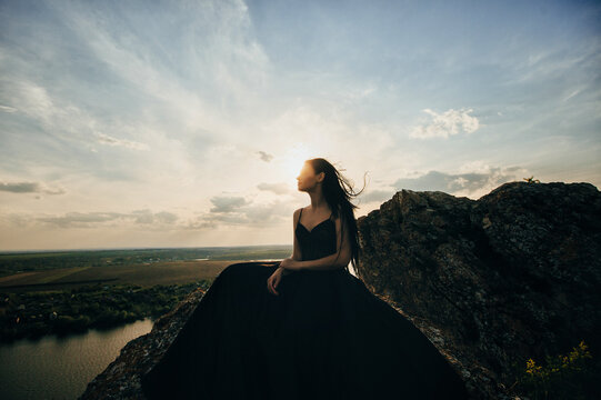 The girl is dressed in a long black dress. Standing on a rock. High above the river. Nice view. Back light. Summer. Dramatic photos.