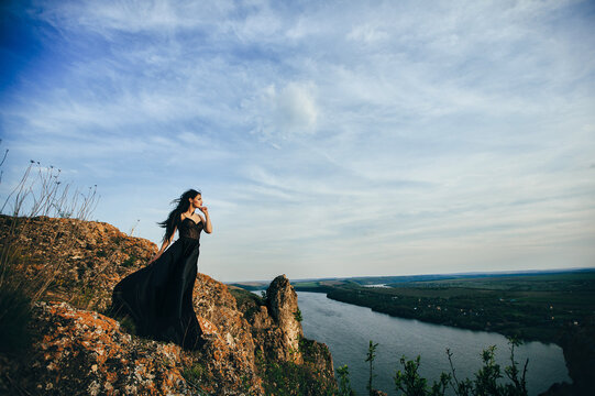 The girl is dressed in a long black dress. Standing on a rock. High above the river. Nice view. Back light. Summer. Dramatic photos.