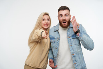 Fototapeta premium Amazed young pretty couple raising emotionally their hands while looking surprisedly at camera with wide eyes opened, standing over white background