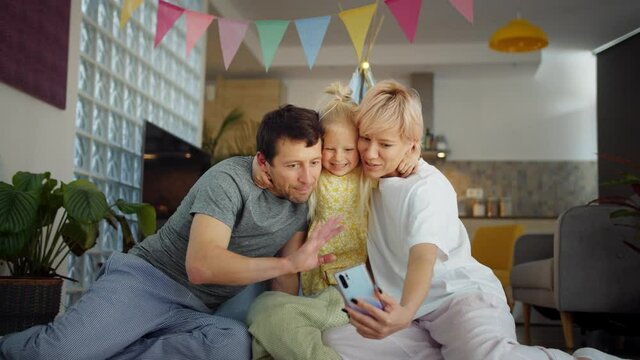 Beautiful Caucasian family making video call online using new application on device, happy blonde mother holding light smartphone and handsome father welcome gesture with little daughter at home