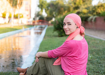 girl with pink cancer scarf sitting on grass happy smiling