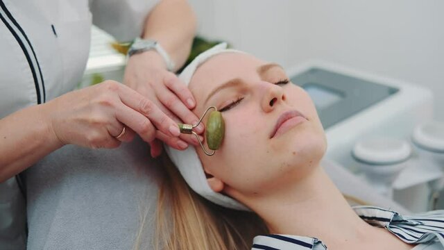 Close-up shot of facial massage with a jade roller on woman's face at beauty salon. Cosmetologist doing procedure for young woman.