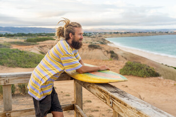 Portrait of Hipster Surfer with dreadlocks and beard looking at the ocean with vintage surfboard .
