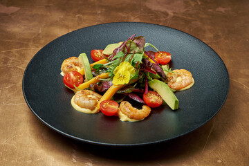 Salad with fried shrimp, arugula, cherry tomatoes, avocado and mango in a black ceramic plate on a copper, metal background. Close up. Seafood salad