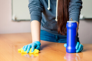 girl in a gray sweater and blue gloves washes a wooden table with a yellow rag.