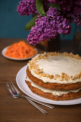 Carrot cake with hazel nuts, grated carrot and bouquet of purple lilac on the wooden table