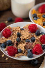 Corn flakes with blueberries and raspberries in white bowl in dark wooden desk.