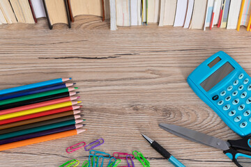 The new school and office supplies lie on a wooden desk top next to a row of books.