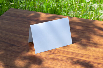 Blank paper table card standing on a wooden table outdoors with blurred green spring backyard background as template for design presentation, event promotion, showcase etc.