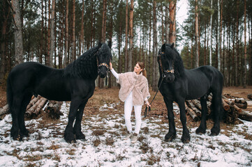 Magnetic young female in grey fur coat and white trousers stands with two big black horses outside
