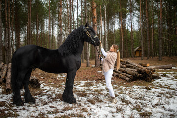 Handsome caucasian woman with fair hair in warm clothes with a beautiful black horse on nature