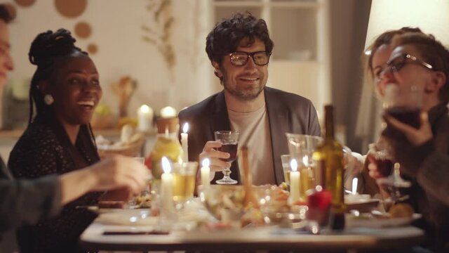 Company of multiethnic male and female friends sitting together at dinner table with candles and food on it, chatting, clinking glasses together and drinking wine while celebrating birthday at home