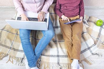 A young woman and a preschooler boy are sitting on a plaid blanket on the floor in the apartment. Mom works on a laptop and her son plays on a tablet nearby, remote work from home, quarantine.