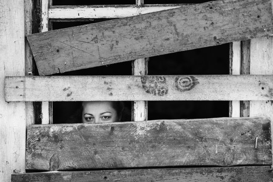 Locked up illegal refugee girl in abroad shelter portrait. Woman slavery. Werid strange unusual person looking through slit in boarded up window in old wooden abandoned ghost house. Social problems.