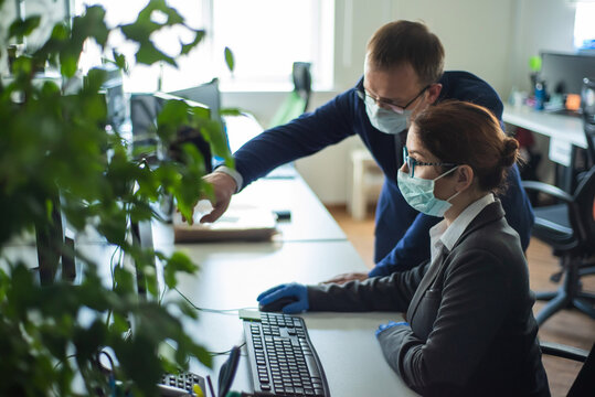 Colleagues work in surgical masks in an open office space communicate at the work desk. A male top manager teaches a female intern how to work at a computer. The boss directs the subordinate.