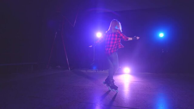 Charming european smiling girl on roller skates performs various turns and elements, skating in abandoned building site at night under pink and blue light colors and smoke in a studio.