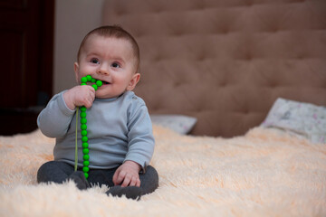 Little boy sits on the bed and smiles. A little boy holds green beads in his mouth.