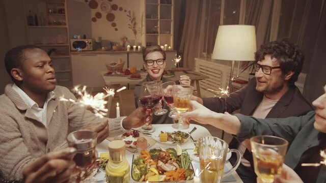 Zoom out shot of happy multiethnic men and women holding sparklers, clinking glasses and drinking wine after a toast while having Christmas dinner party at home