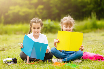 two cute multicultural schoolgirls sitting on lawn under tree and reading book together