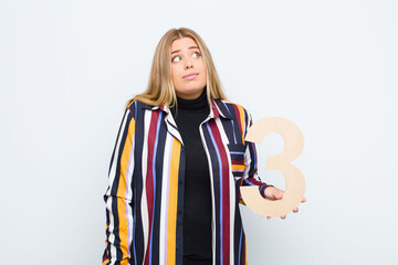 young pretty blonde woman confused, doubtful, thinking, holding a number 3.