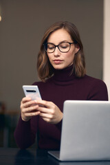 Young beautiful woman with glasses, sitting in front of her desk while using her smartphone