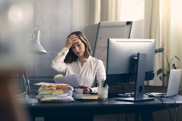 Very stressed business woman sitting in front of her computer looking at a large pile of paperwork, while holding a hand at her forehead 