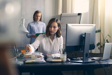 Busy female CEO looking through a large pile of paper applications at her modern office desk while her employee is walking in the background. 