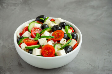 Greek salad of fresh juicy vegetables, feta cheese, herbs and olives in a white bowl on a dark concrete background. Healthy food. Horizontal orientation. Copy space.