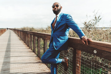 A handsome brutal bald bearded African man in sunglasses and a blue fashionable costume with a tie...
