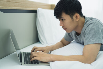 Young Asian man working from home by using computer laptop notebook which lie on bed in bed room which felling be serious. Corona virus, COVID-19 Pandemic new normal life at home Concept.