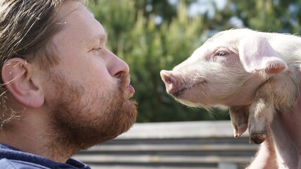 A bearded man holds a pig in his hands and kisses him.