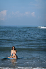 Happy brunette woman sitting on blue surfboard in the ocean. Surfer girl having fun inside ocean during the summer in holiday and activity concept with copy space