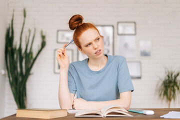 Thoughtful redhead young woman student is making an outline from textbook in workbook at desk at home office.