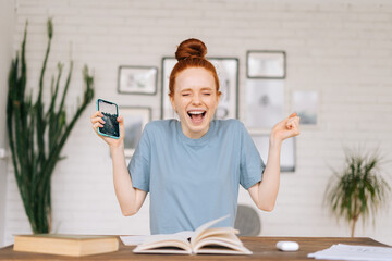 Happy redhead young woman is feeling euphoric reading good amazing news on cell phone while sitting...