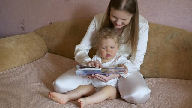 Beautiful attractive mother with a cute baby sit at home on the couch and have fun looking at the picture book.