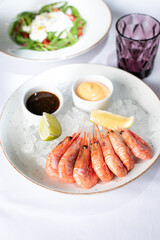 Fresh seafood in a restaurant. Shrimps on a white plate with ice. White tablecloth. Sauces, llama and lemon pussies. Nearby is a glass cup. A plate with salad is visible.