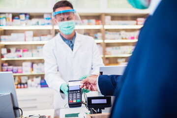 Professional pharmacist with protective mask and face shield on his face  working with customer in modern drugstore.