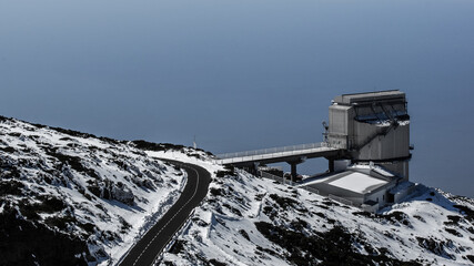 Obraz premium A abstrakt looking building of the observatory on the Canary Island La Palma. A road leads to the building. The ground is covered with snow and in the background the Atlantic Ocean is visible.